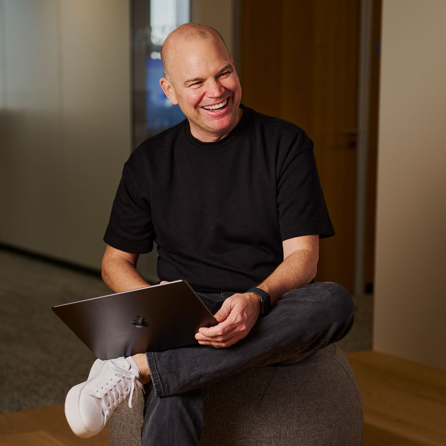 Der Lexware-Geschäftsführer Christian Steiger sitzt lächelnd mit einem Laptop auf dem Schoß auf einem Stuhl. Er trägt ein schwarzes T-Shirt.