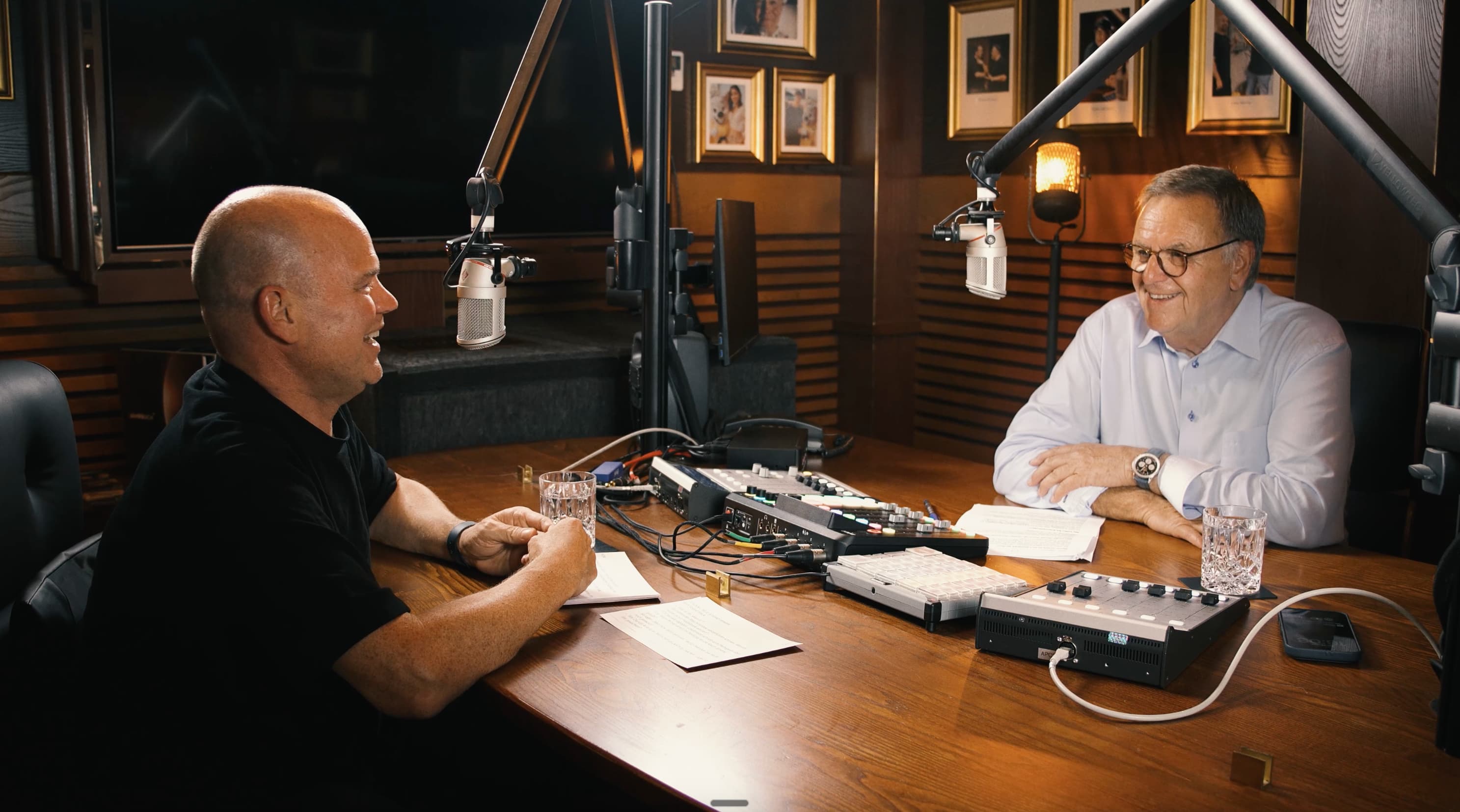 Christian Steiger und Roland Mack unterhalten sich lächelnd bei einem Podcast mit Mikrofonen und Mischpult am Holztisch.