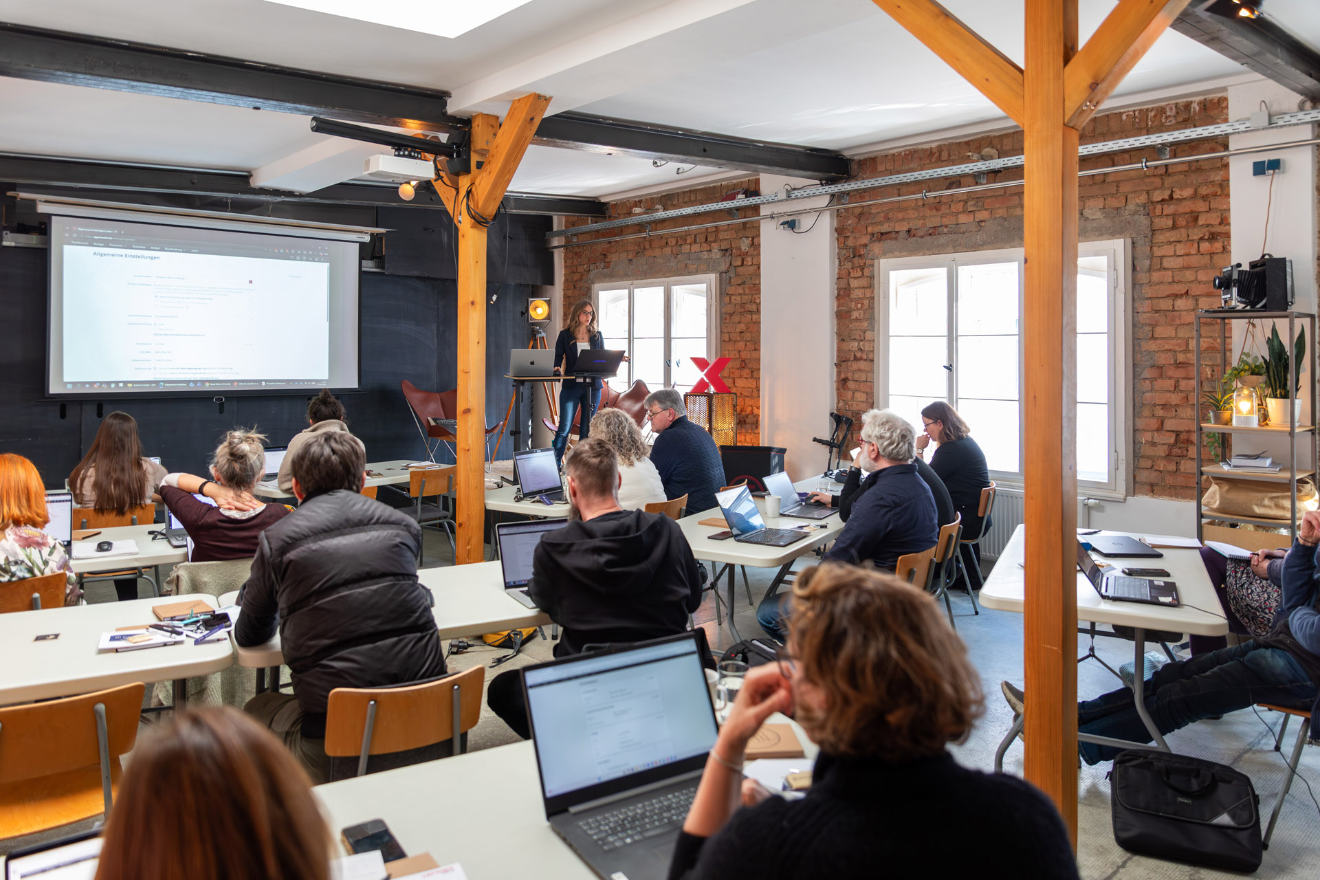 Teilnehmende beim Lexware-Office-Coaching mit Laptops in einem Backstein-Loft, vorne eine Trainerin am Stehpult vor der Leinwand.
