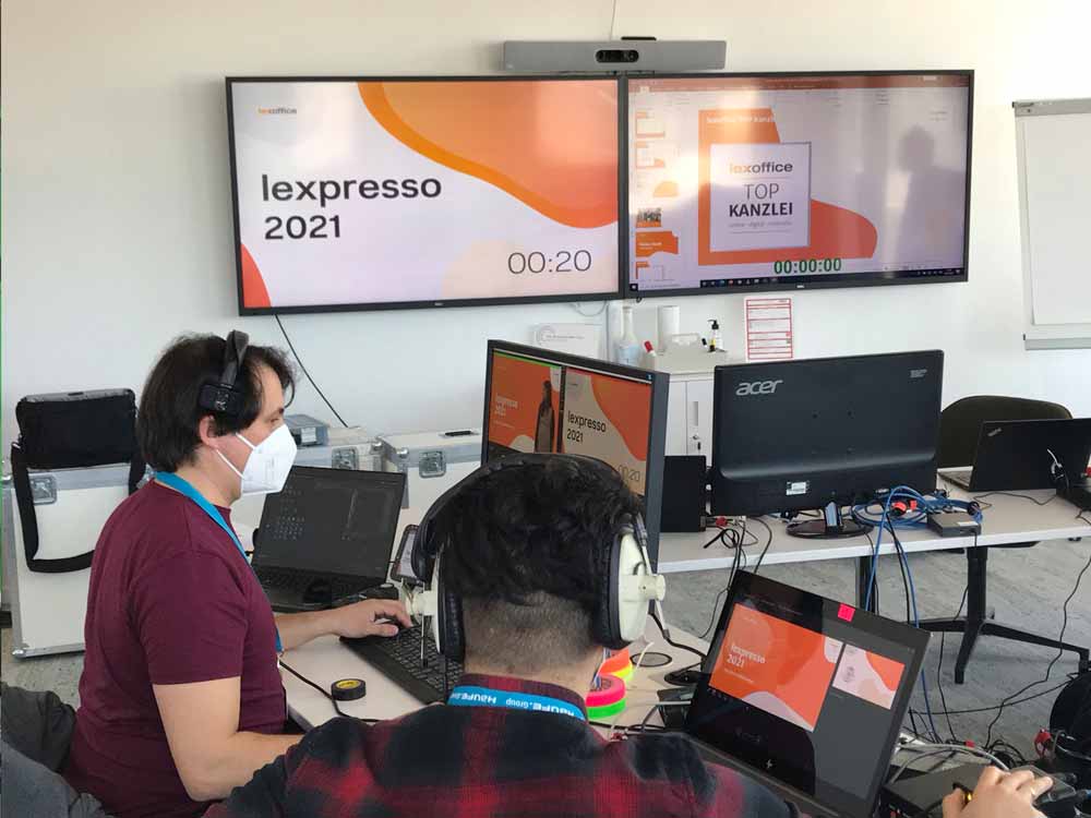 Blick auf zwei Bildschirme im Regieraum: einer zeigt die Startgrafik der lexpresso 2021, der andere eine Folie mit der Aufschrift lexoffice TOP KANZLEI, davor sitzen zwei Personen mit Kopfhörern und Maske an Laptops