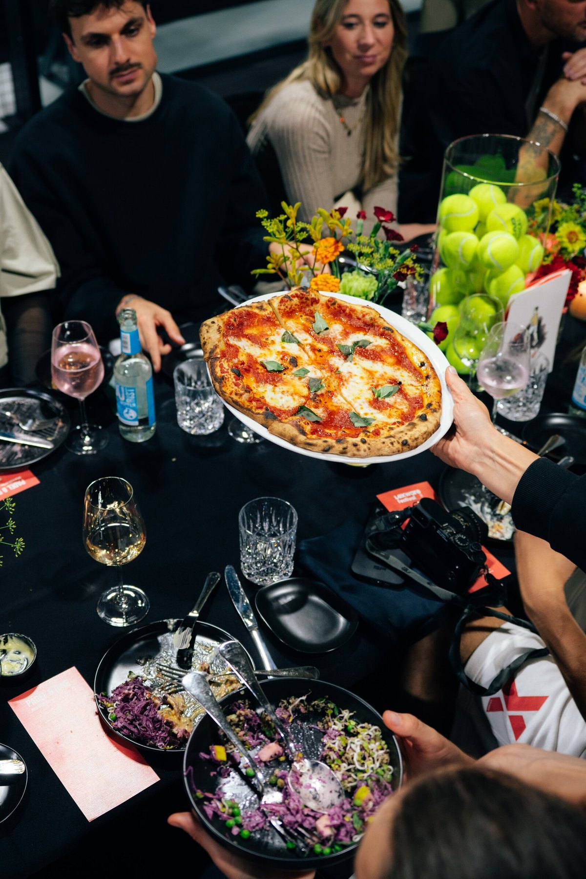 Frisch gebackene Pizza wird über die gedeckte Tafel gereicht, daneben Tennisbälle und Blumen.