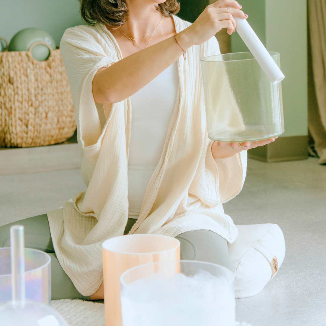 A person sitting cross-legged on a white cushion on the floor, holding a transparent crystal singing bowl in one hand and a white mallet in the other, surrounded by other crystal singing bowls, in a calm, softly lit room.