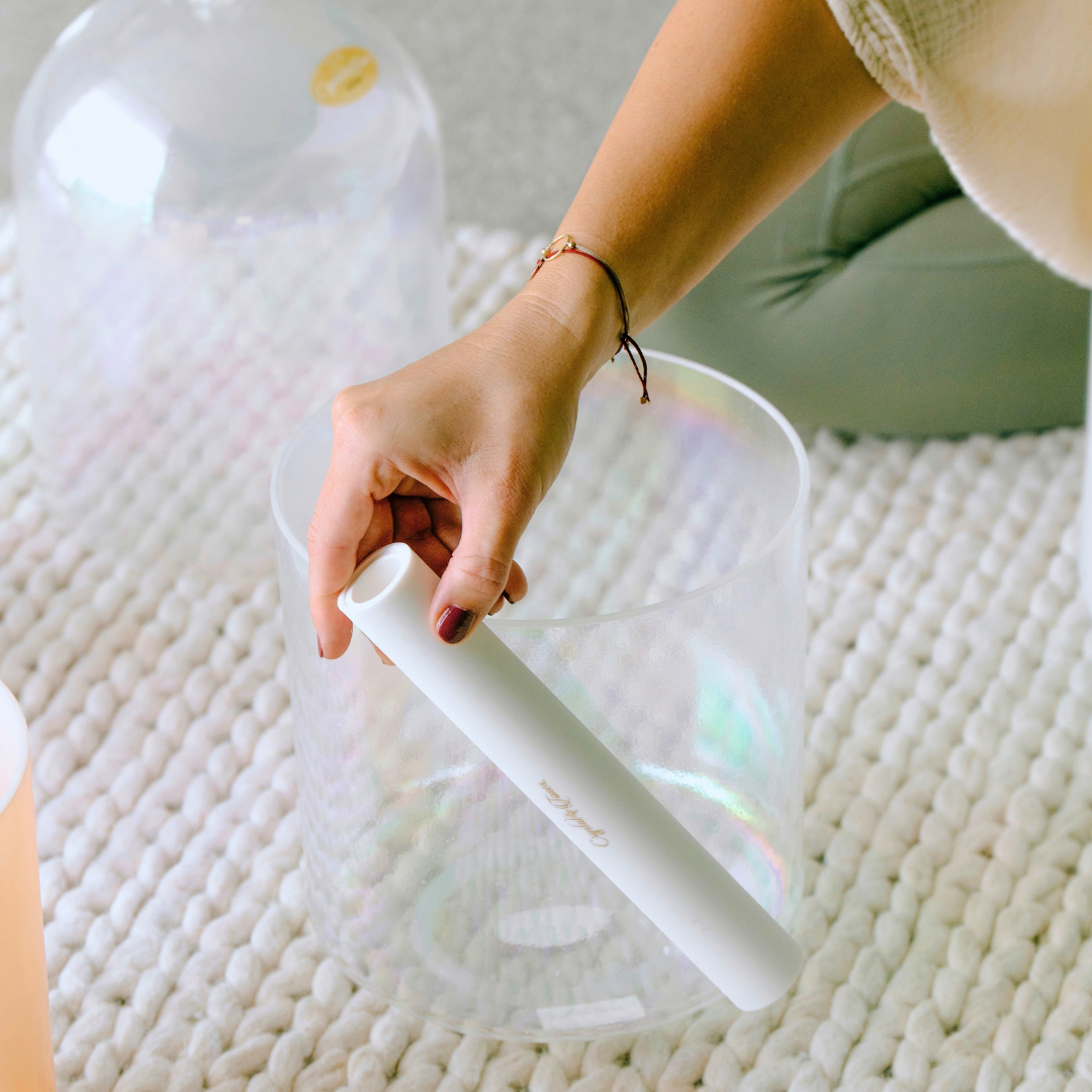A person holding a white cylindrical mallet above a translucent crystal singing bowl on a textured white rug, with a large glass vessel in the background.