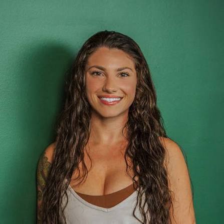 A woman with long, curly brown hair smiling against a solid green background, wearing a gray tank top over a brown sports bra.