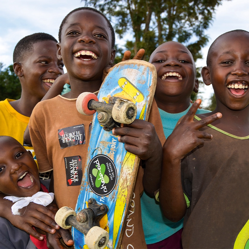 Gruppe lächelnder Jungen im Freien, einer hält ein abgenutztes blaues Skateboard mit Aufklebern.