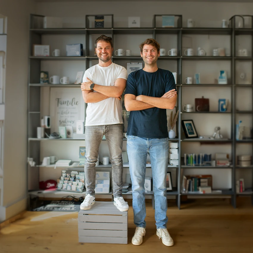 Benedikt und Leo stehen vor einem Regal, lächeln in die Kamera und halten ihre Arme verschränkt vor dem Körper.