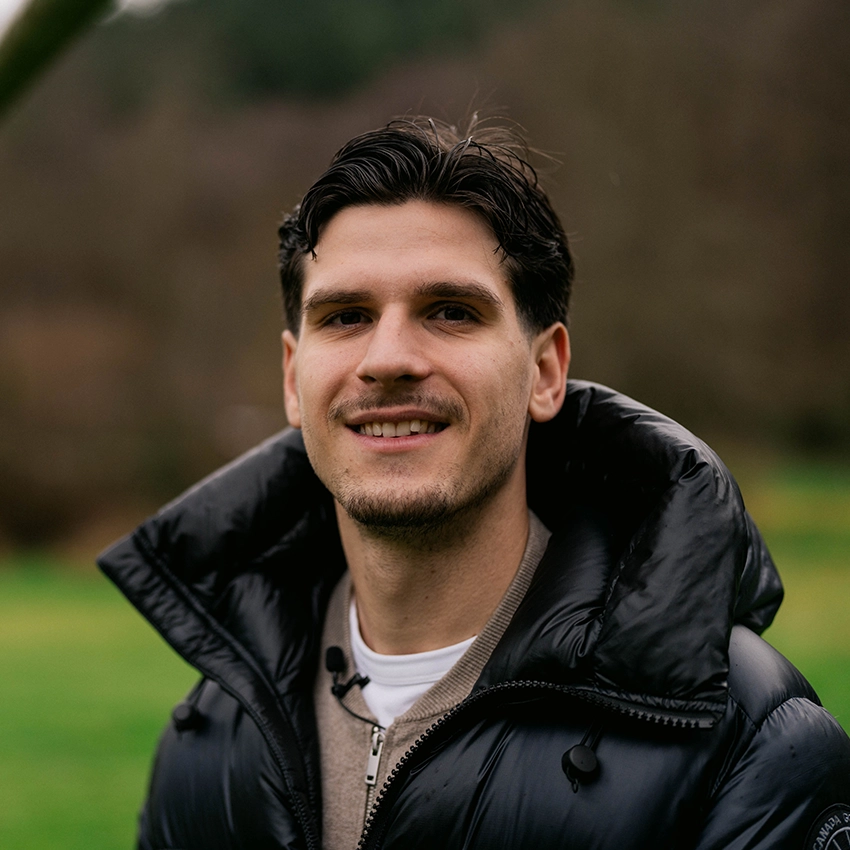 Igor Matanovic steht im Freien, trägt eine schwarze Daunenjacke, einen beigen Pullover und ein weißes Hemd, lächelt in die Kamera.