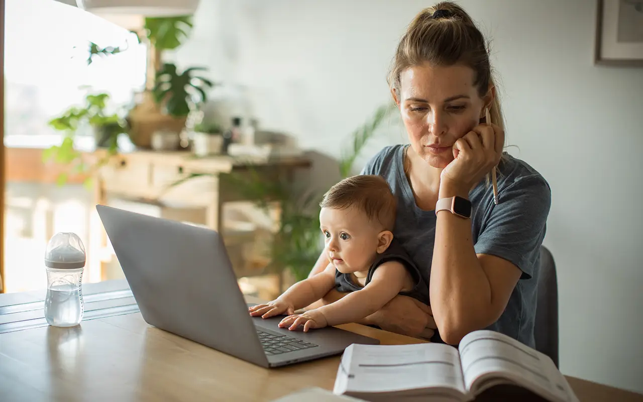 Frau sitzt mit Baby auf dem Schoß vor Laptop und schaut in ein Buch - Beitragsbild von Lexware Tell Your Story