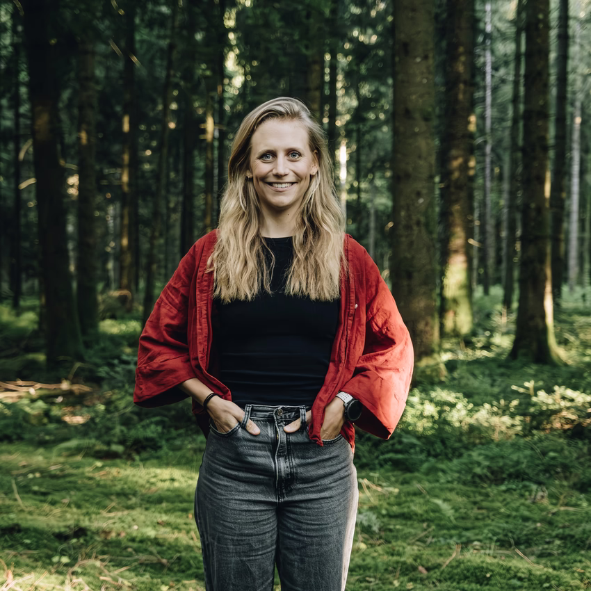 Wiebke steht lächelnd mit den Händen in den Hosentaschen in einem grünen Wald, sie trägt eine rote Jacke über schwarzem Shirt.