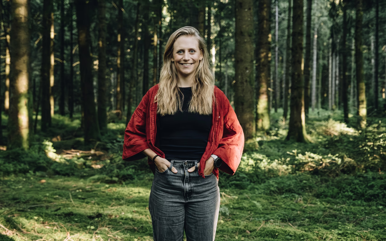 Wiebke steht lächelnd mit den Händen in den Hosentaschen in einem grünen Wald, sie trägt eine rote Jacke über schwarzem Shirt.