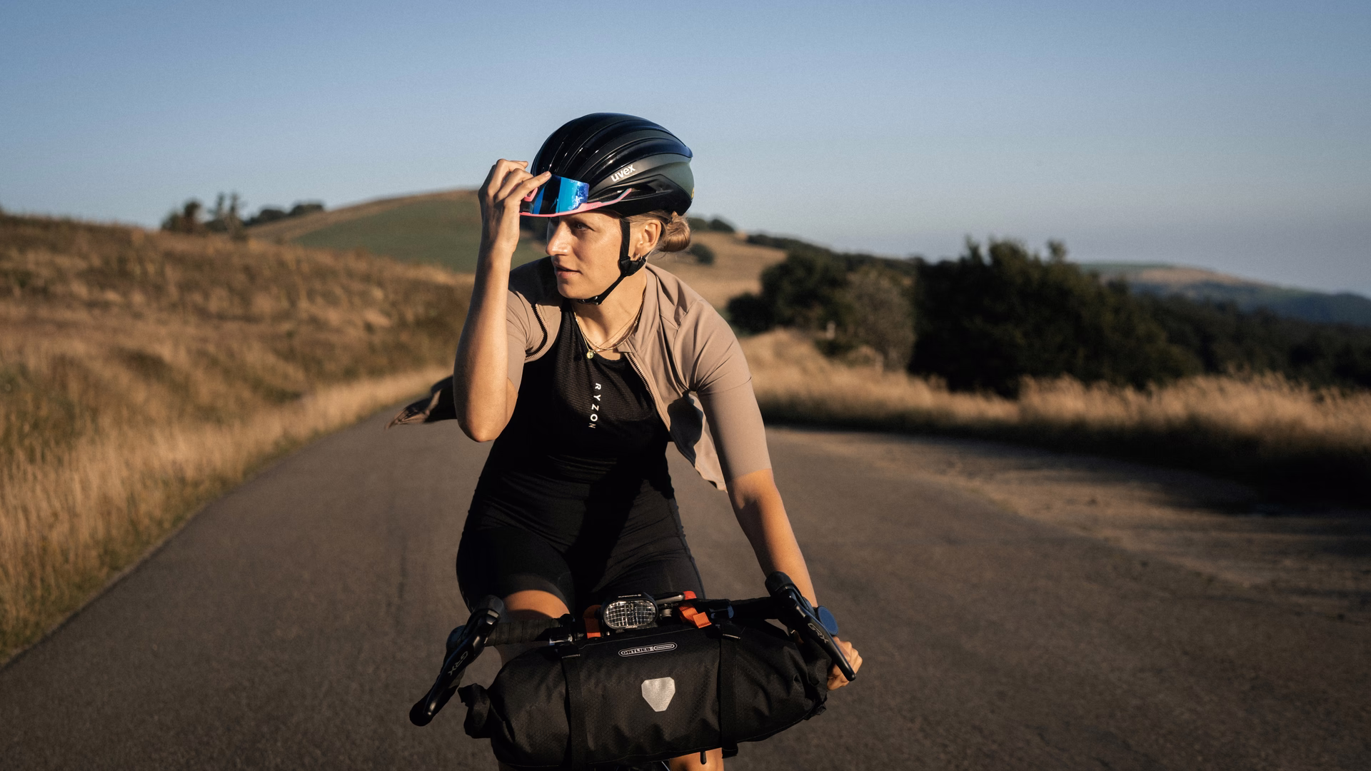 Radsportlerin Wiebke fährt bei goldenem Abendlicht auf einer Landstraße und schiebt ihre verspiegelte Sonnenbrille hoch, im Hintergrund sanfte Hügel.