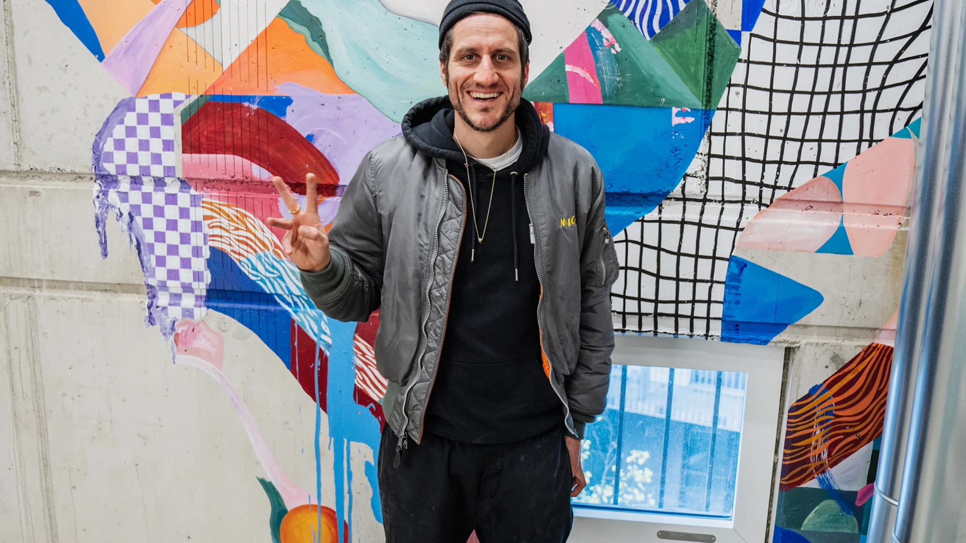 Micha Fritz steht vor einer bunt bemalten Wand und formt das Peace-Zeichen mit seinen Fingern.