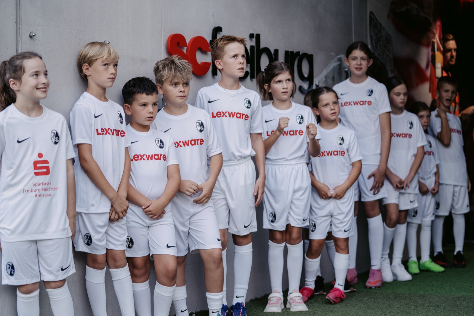 Kinder in weißen Lexware-Trikots vor der "scfreiburg"-Wand im Tunnel, zeigt genau den beschriebenen Moment vorm Einlaufen