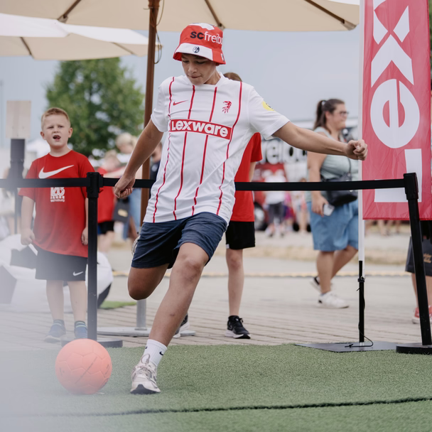 Jugendlicher im SC-Freiburg-Trikot mit Lexware-Logo schießt in der Fanzone einen Ball, im Hintergrund Besuchende und eine rote Lexware-Fahne.