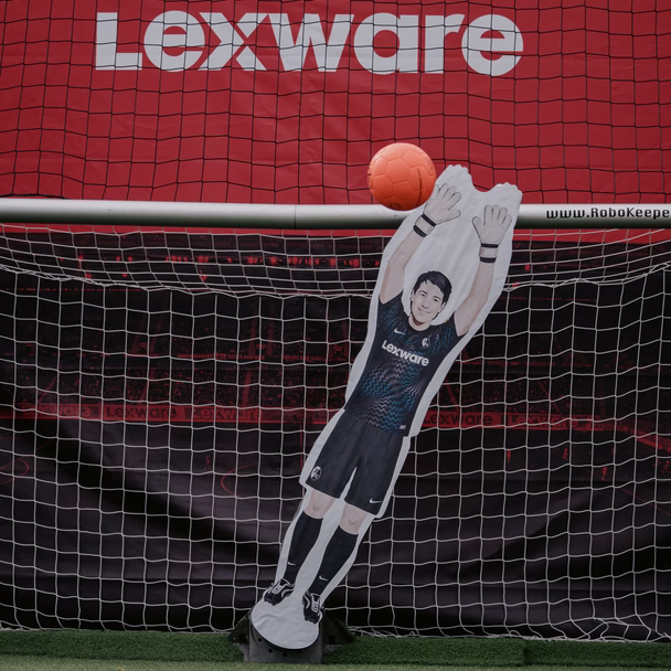 Pappaufsteller eines Torhüters im Lexware-Trikot streckt sich nach einem orangefarbenen Ball, dahinter ein Fußballtor mit rotem Lexware-Banner.
