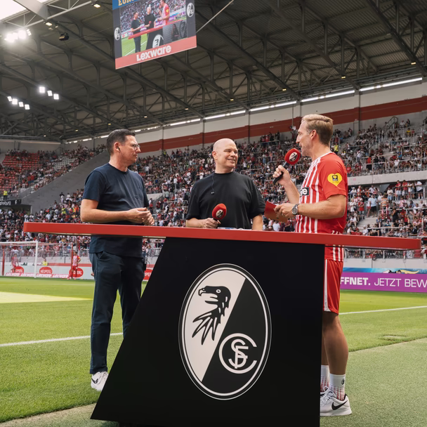 Christian Steiger im Gespräch an einem Stehtisch mit SC-Freiburg-Wappen am Spielfeldrand, im Hintergrund die gefüllten Ränge des Stadions.