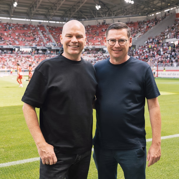 Zwei Männer stehen lächelnd nebeneinander am Spielfeldrand, im Hintergrund das Spielfeld und die Zuschauerränge.