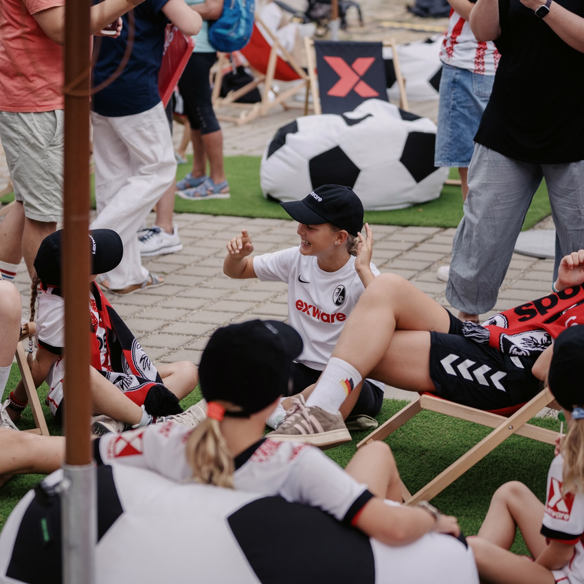 Kinder und Jugendliche in SC-Freiburg-Trikots mit Lexware-Logo sitzen in Liegestühlen und auf dem Rasen der Fanzone, daneben ein großer Sitzsack in Fußballoptik.