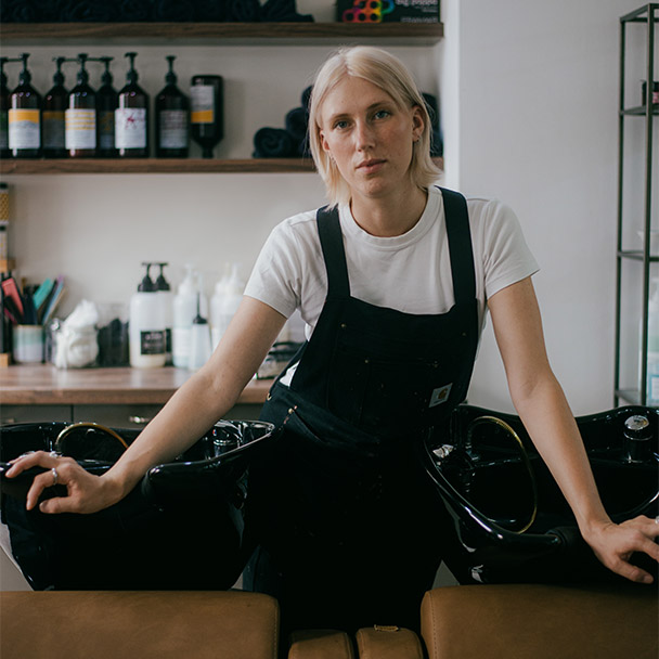 Franka Bähr lehnt an den Waschbecken ihres Salons, hinter ihr ein Regal mit Pflegeprodukten.