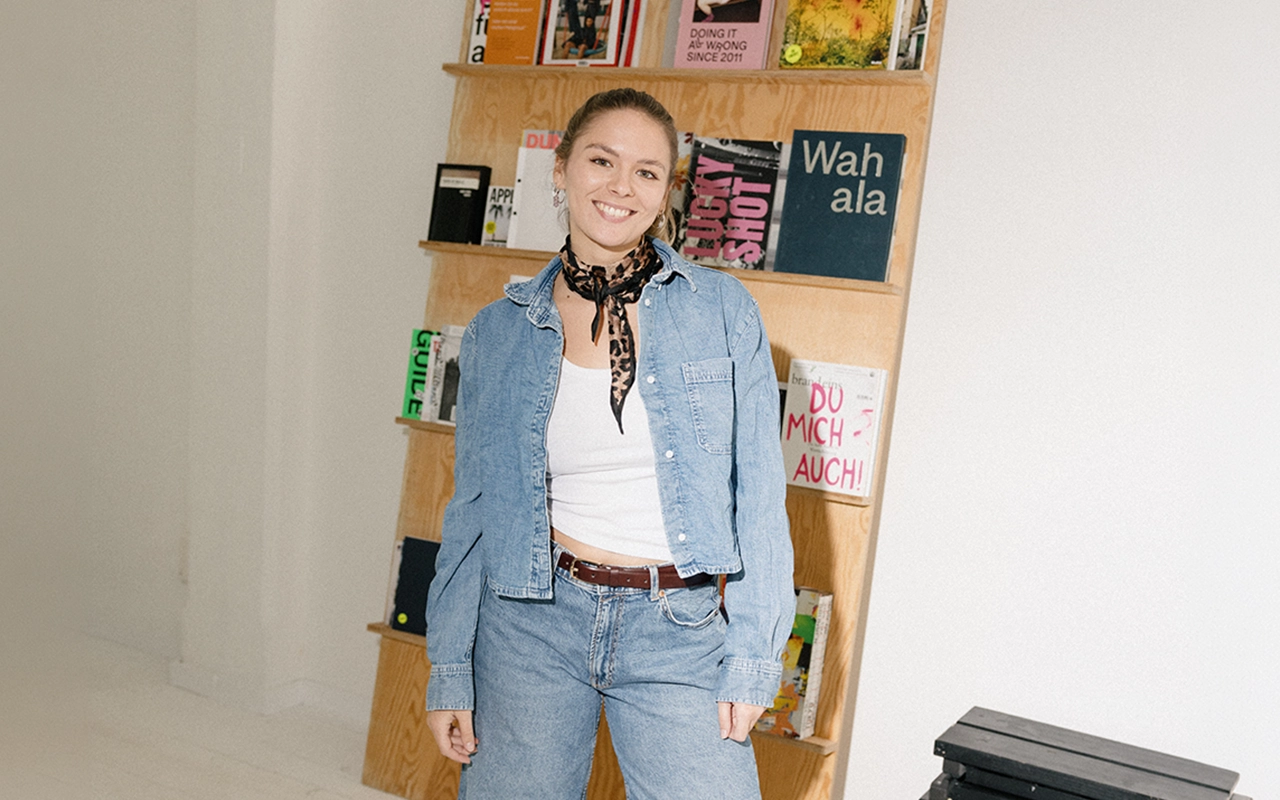 Francesca Amann steht lächelnd vor einem Holzregal voller Magazine, in Jeansjacke und mit Leopardentuch.