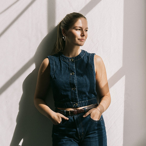 Francesca Amann lehnt in dunkler Jeansweste an einer weißen Wand, auf der Schatten von Sonnenlicht liegen.