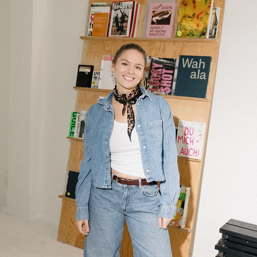 Francesca Amann steht lächelnd vor einem Holzregal voller Magazine, in Jeansjacke und mit Leopardentuch.