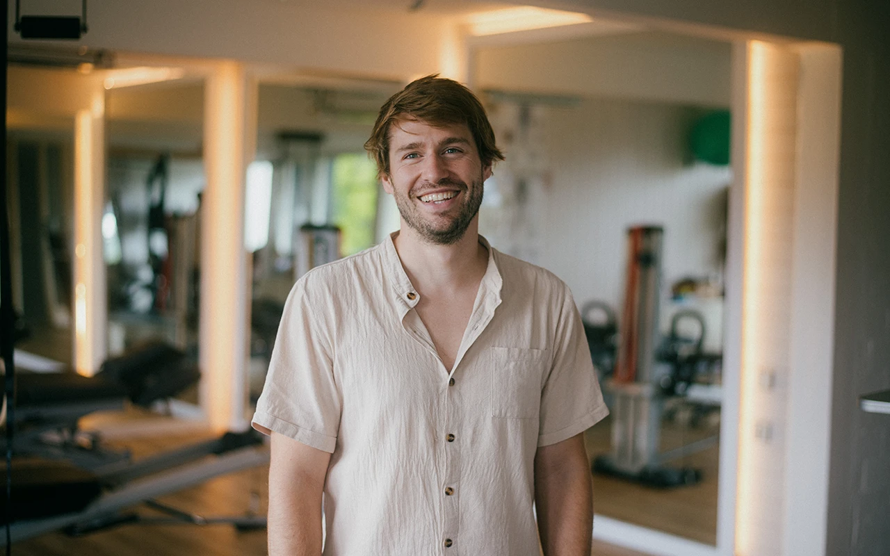 Physiotherapeut Tom Gabler steht lächelnd in seinem Praxisraum, hinter ihm Trainingsgeräte und beleuchtete Wandnischen.
