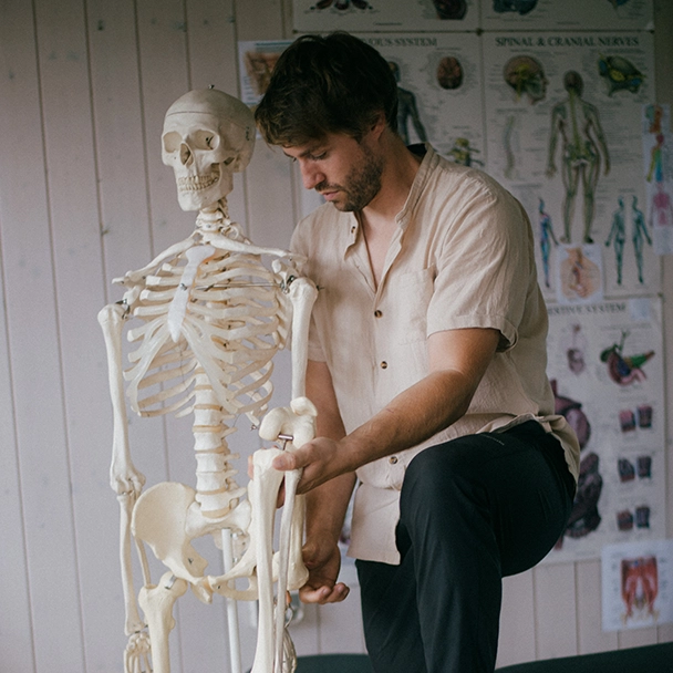  Tom Gabler beugt sich zu einem Skelettmodell und hält dessen Kniegelenk, im Hintergrund Anatomie-Poster und Behandlungsliege.