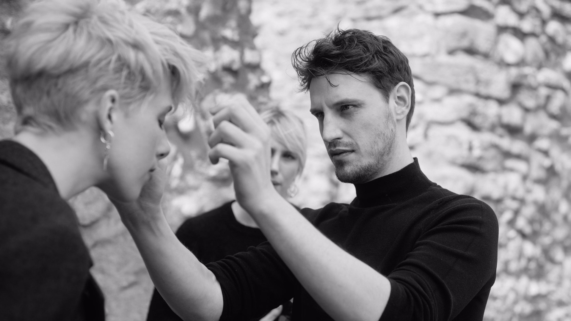 Friseur im schwarzen Rollkragenpullover richtet konzentriert das kurze blonde Haar einer Kundin, Schwarzweißfoto vor einer Natursteinmauer.