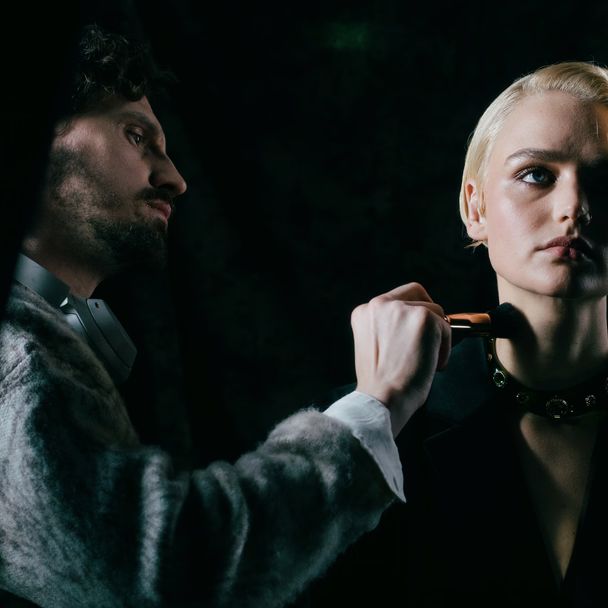 Stylist setzt einer Kundin mit blondem Kurzhaarschnitt einen Make-up-Pinsel an Kinn und Hals, dramatisches Licht vor dunklem Hintergrund.