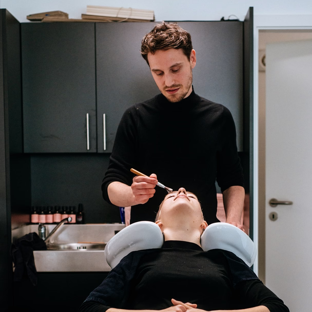 Friseur trägt einer liegenden Kundin am Waschbecken mit einem Pinsel Farbe am Haaransatz auf.