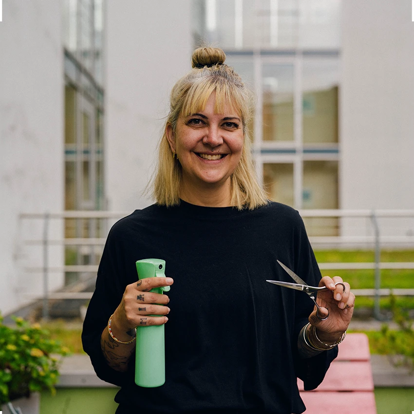 Steffi Schöne lächelt in die Kamera und hält eine grüne Sprühflasche und eine Friseurschere in den Händen.