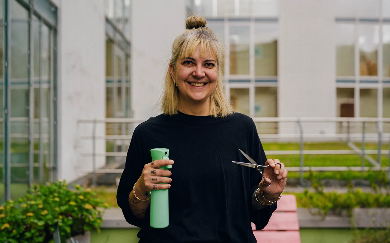 Steffi Schöne lächelt in die Kamera und hält eine grüne Sprühflasche und eine Friseurschere in den Händen.