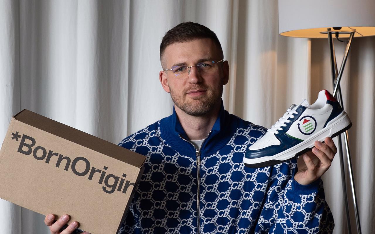 Melvin Lamberty sitzt auf einem Sessel und hält einen Born-Originals-Karton sowie einen weiß-blauen Sneaker in die Kamera.