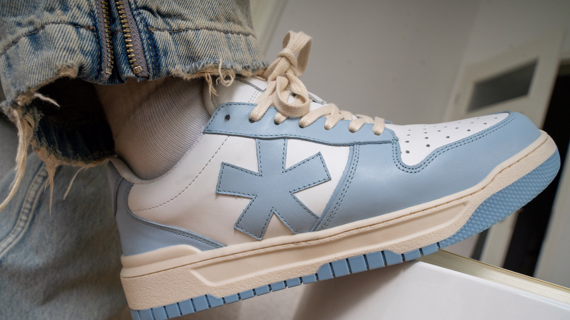 Hellblau-weißer Born-Originals-Sneaker mit Stern-Logo an einem Fuß, das Bein über eine weiße Kante gestreckt.