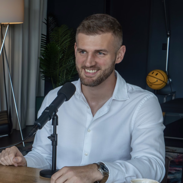 Melvin Lamberty sitzt lachend vor einem Mikrofon an einem Holztisch bei einer Podcastaufnahme.