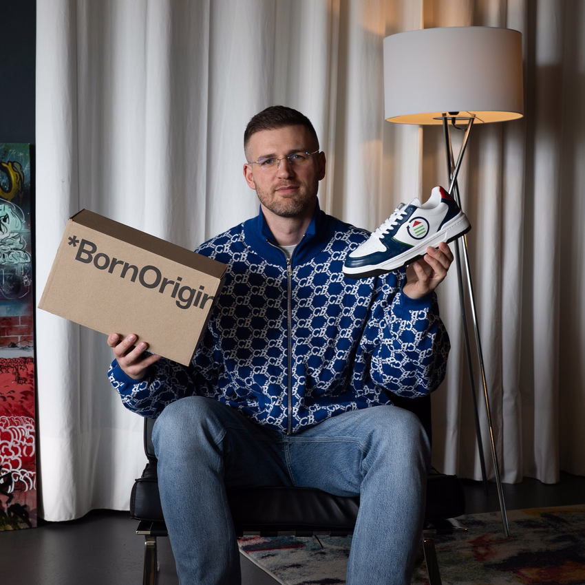 Melvin Lamberty sitzt auf einem Sessel und hält einen Born-Originals-Karton sowie einen weiß-blauen Sneaker in die Kamera.