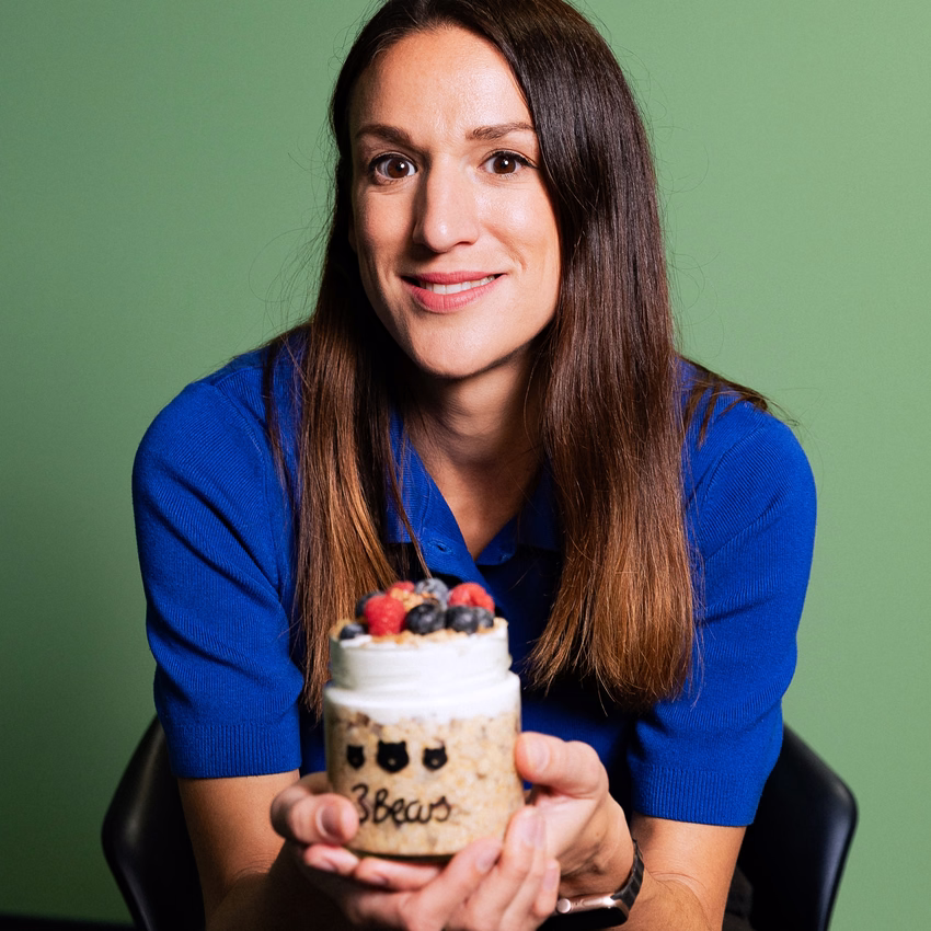 Caroline Nichols hält ein Glas Overnight Oats mit Beeren und Joghurt in die Kamera, im Hintergrund eine grüne Wand.