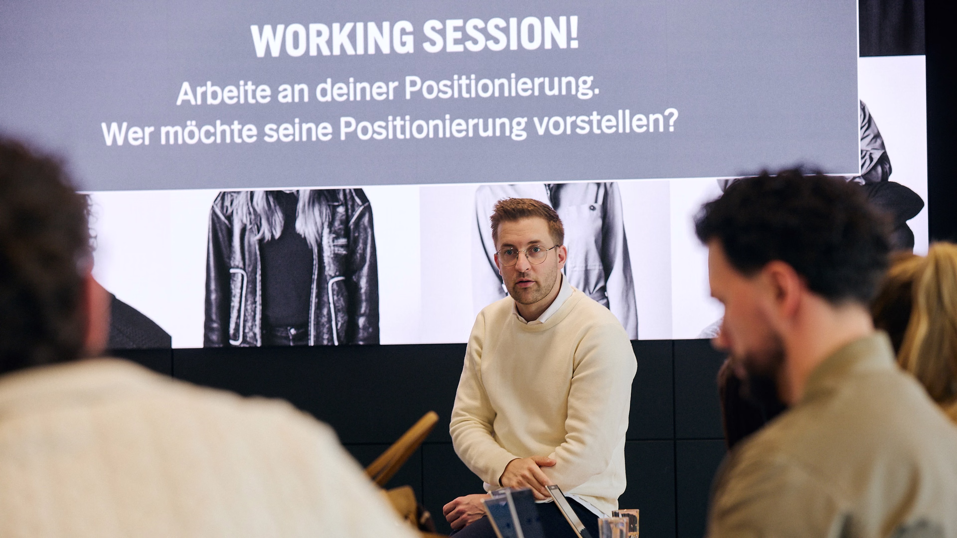 Lukas Weisser sitzt in einem Workshop vor einer Leinwand mit der Aufschrift „Working Session!“, im Vordergrund Teilnehmende am Tisch.
