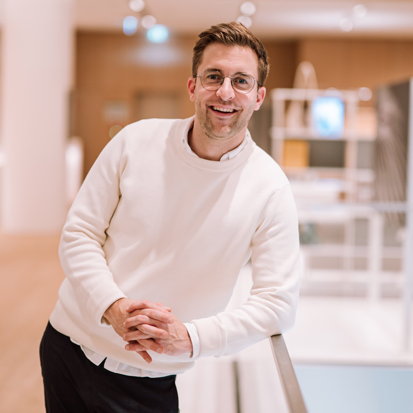 Lukas Weisser lehnt lächelnd an einem Geländer in einem hellen Bürogebäude, er trägt einen weißen Pullover.