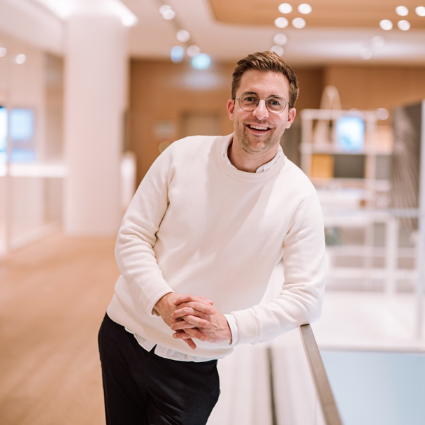 Lukas Weisser lehnt lachend an einem Geländer in einem hellen Bürogebäude, er trägt einen weißen Pullover.