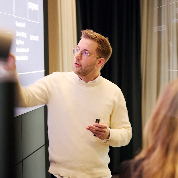 Lukas Weisser steht vor einer Leinwand mit Workshop-Agenda und zeigt auf einen Programmpunkt, in der Hand einen Presenter.