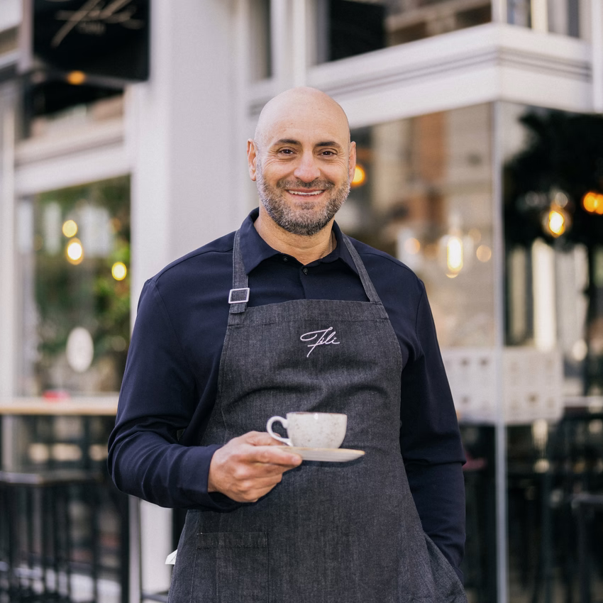 Filipos Klein steht mit Schürze und Kaffeetasse vor einem seiner Fili Cafés.
