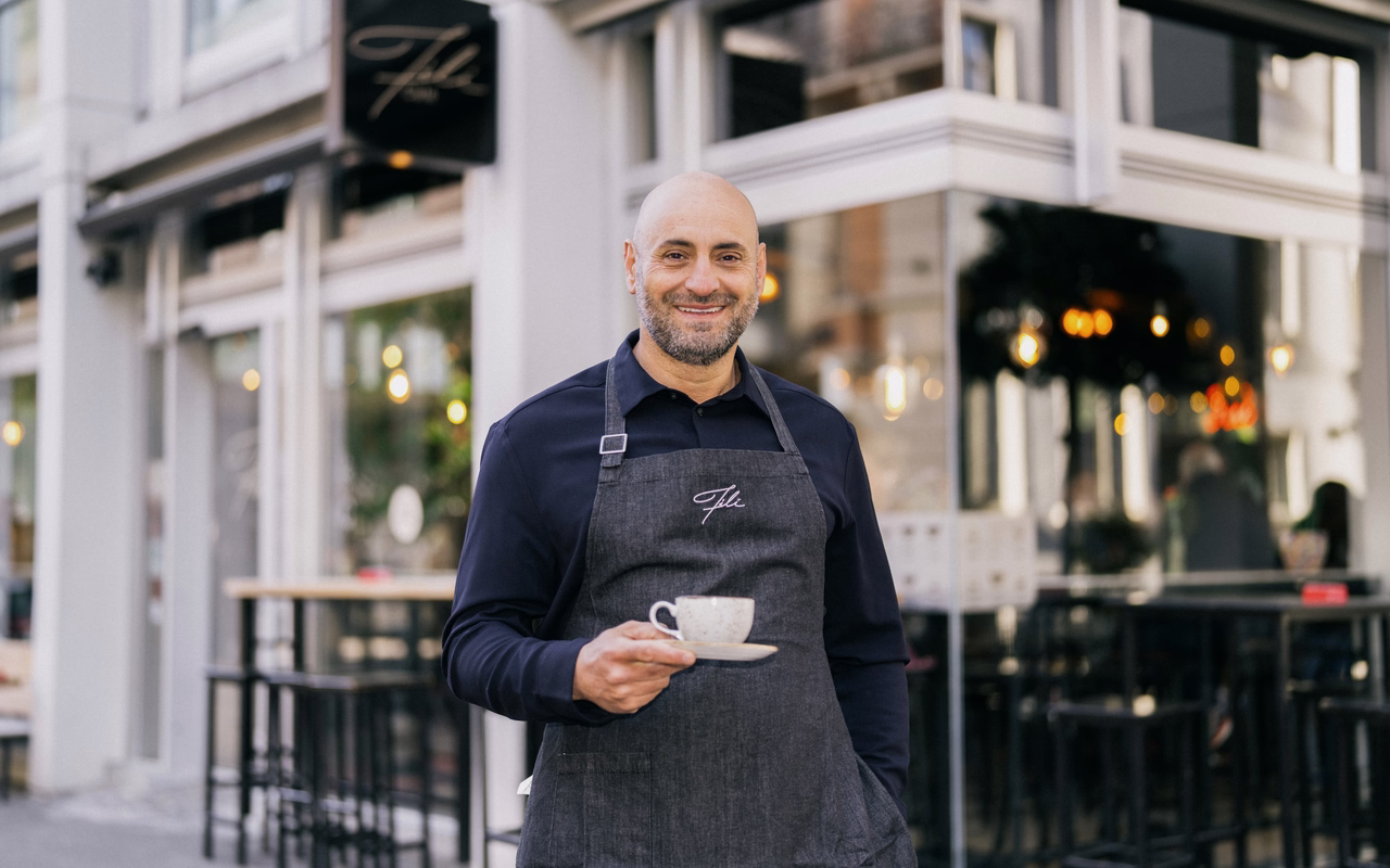 Filipos Klein steht mit Schürze und Kaffeetasse vor einem seiner Fili Cafés.