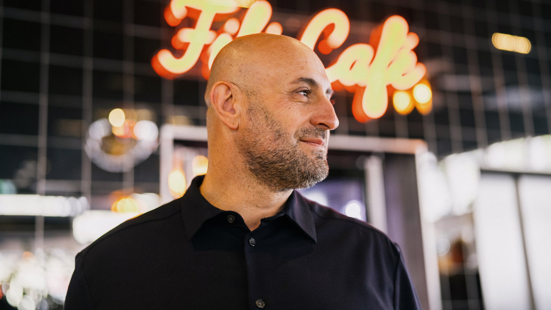 Filipos Klein vor dem leuchtenden Schriftzug „Fili Café“, den Blick zur Seite gerichtet.