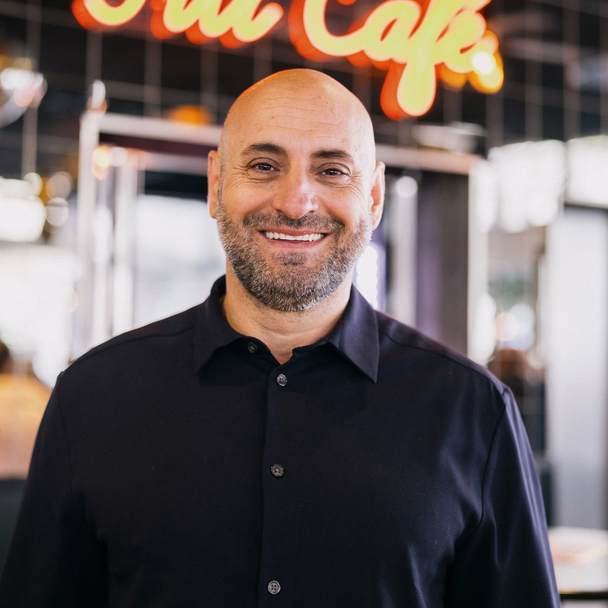 Filipos Klein lächelt in die Kamera, hinter ihm leuchtet der Schriftzug „Fili Café“.