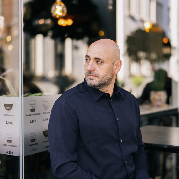 Filipos Klein sitzt mit verschränkten Händen am Fenster eines Fili Cafés und schaut zur Seite.