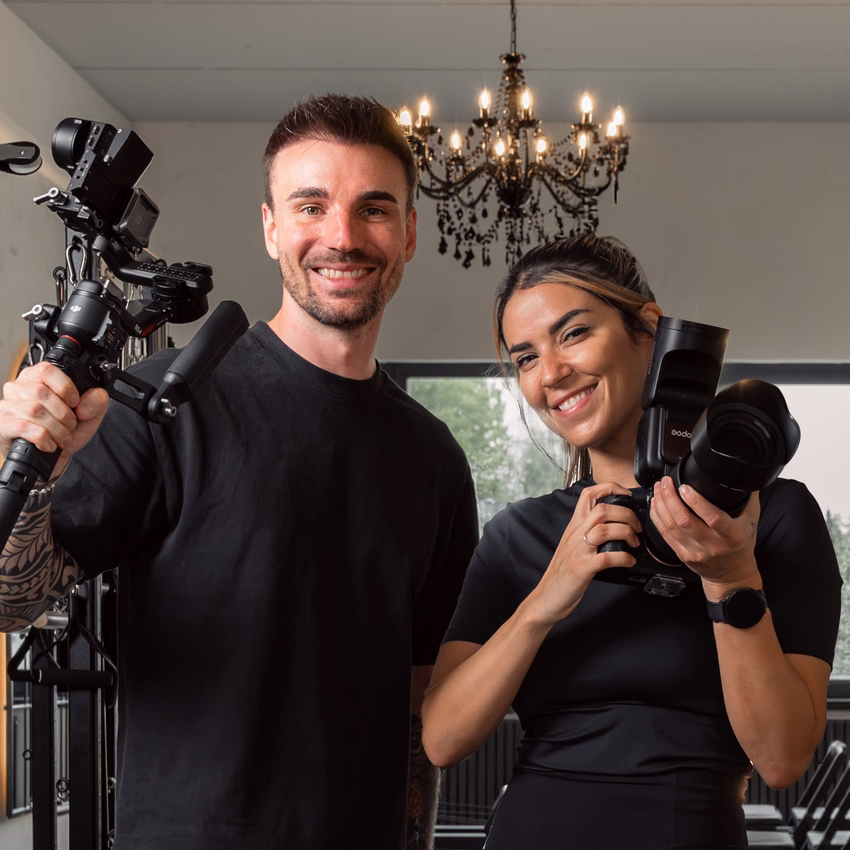 Sören Jeßner mit Gimbal-Kamera und Nathalie Naceur mit Fotokamera lachen in einem Pilates-Studio unter einem Kronleuchter.