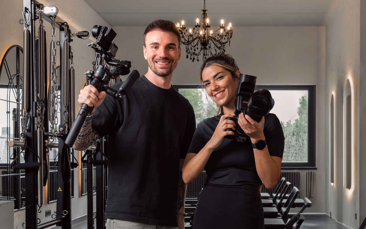 Sören Jeßner und Nathalie Naceur stehen mit Gimbal-Kamera und Fotokamera in einem Pilates-Studio mit Kronleuchter und lachen in die Kamera.