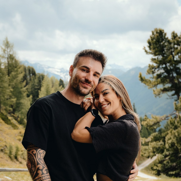 Sören Jeßner und Nathalie Naceur stehen Arm in Arm auf einer Almwiese vor Bergen und Nadelbäumen.